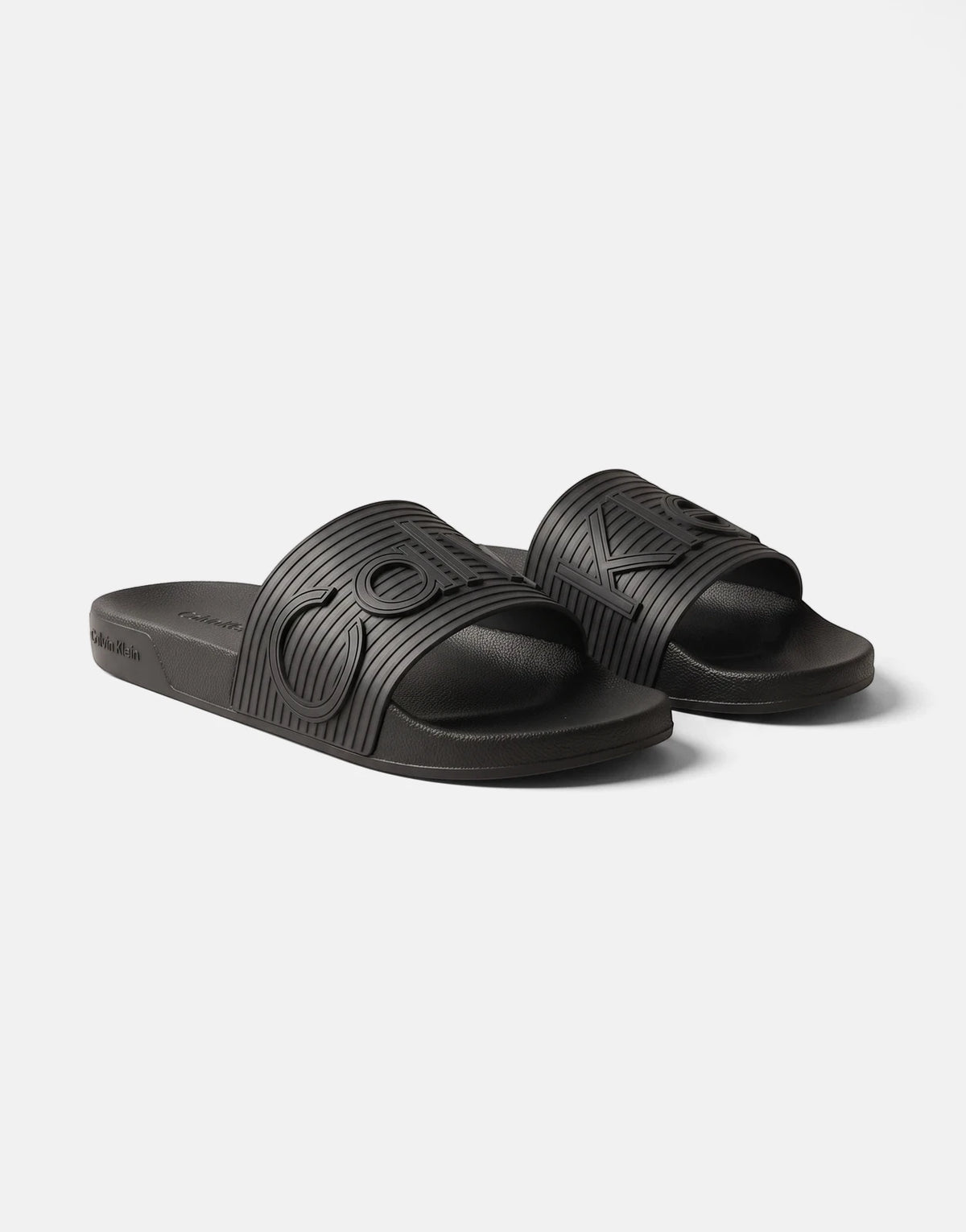 Two black Calvin Klein Essential Texture slides with embossed logos, made of textured rubber, presented at a slight angle.