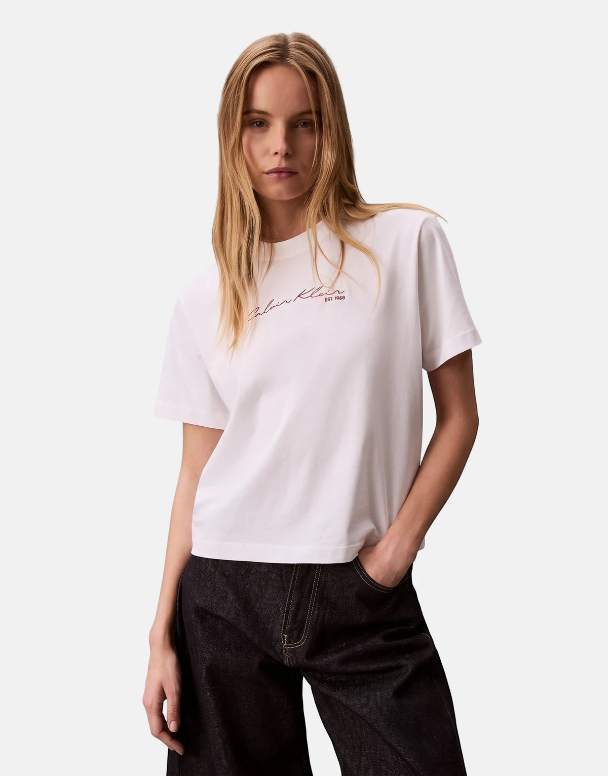 A female model poses wearing a white Calvin Klein Graphic Boxy T-Shirt with a red script logo, paired with dark denim jeans.