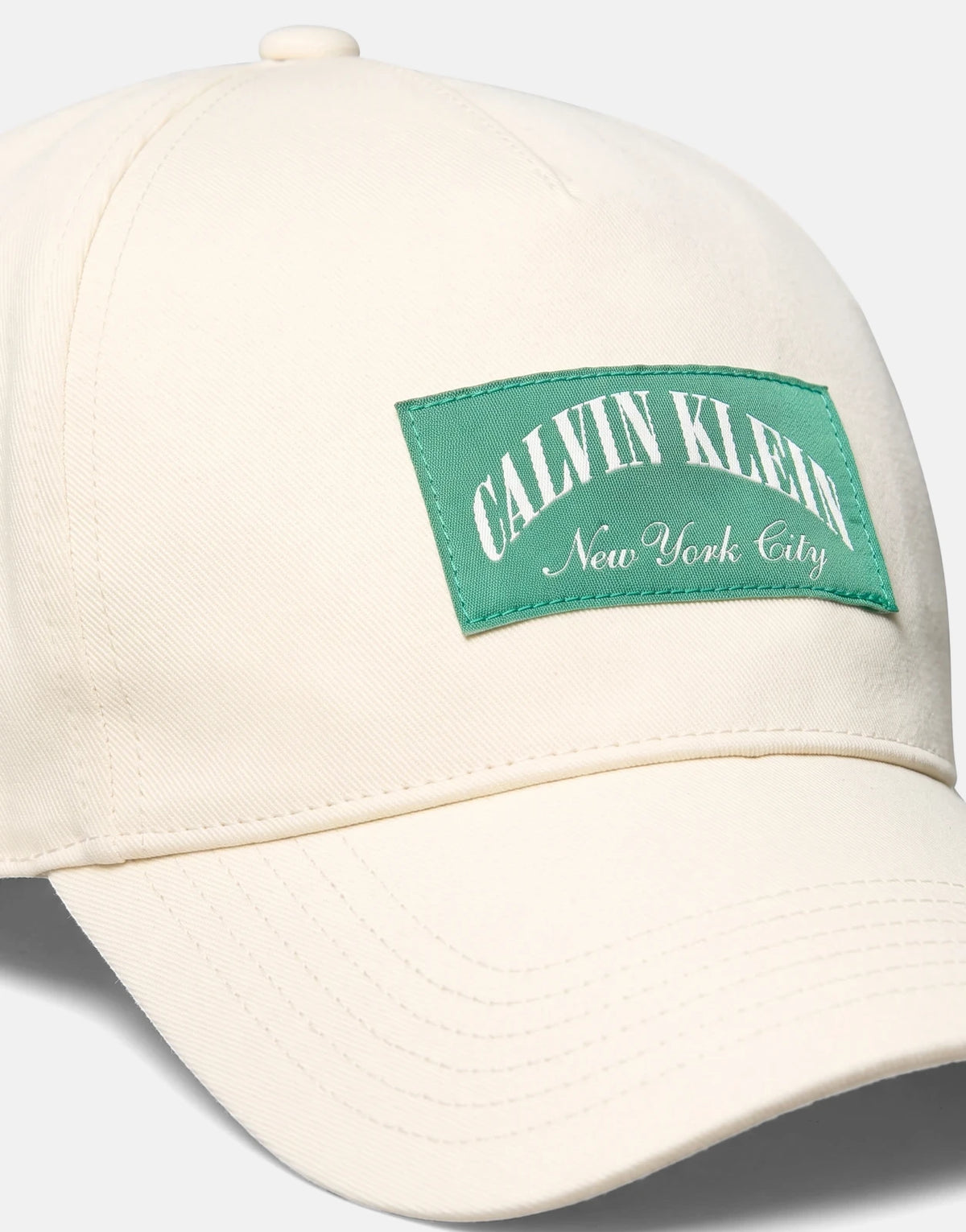 Close-up of a Calvin Klein Heritage Crest off-white pitch cap featuring a green embroidered logo patch on the front.