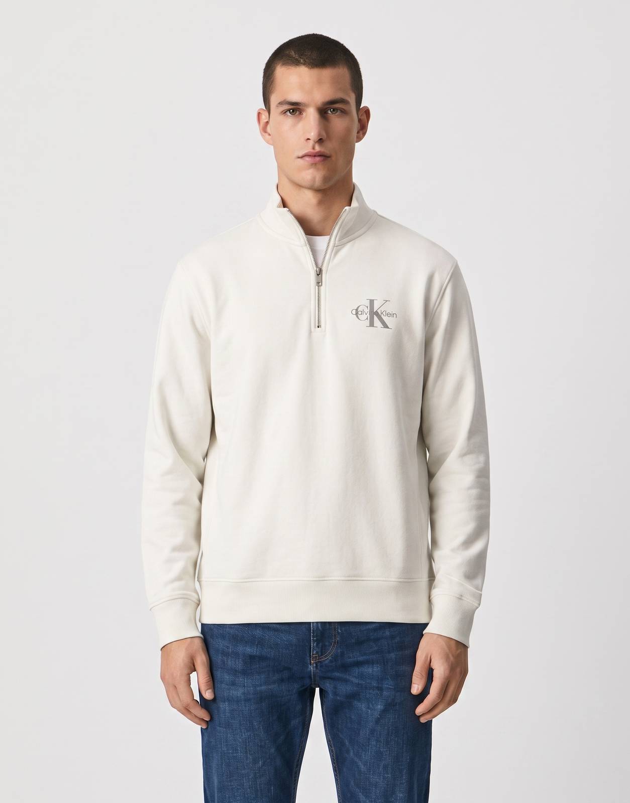 A male model wears a Calvin Klein Micro ML Quarter Zip sweatshirt in off-white, paired with blue jeans and white sneakers.