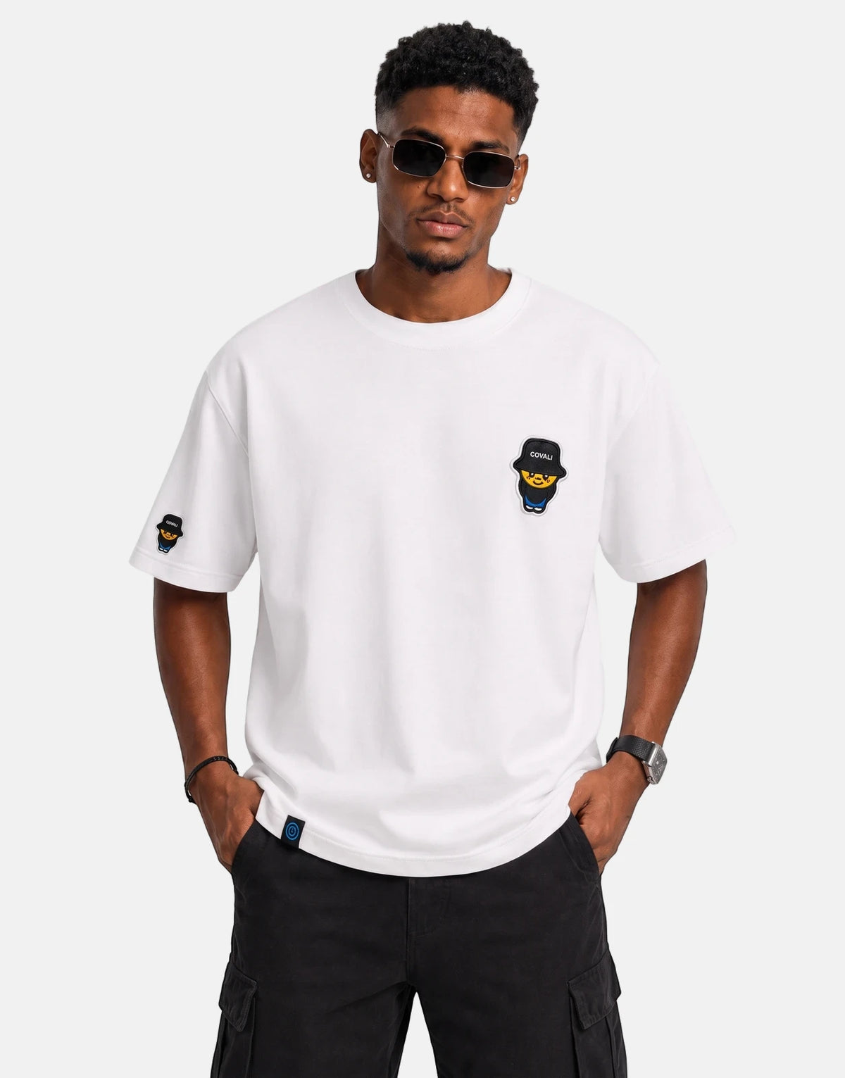 A male model wears a white Covali Tipo t-shirt with a character logo, black cargo pants, and sunglasses in a studio shot.
