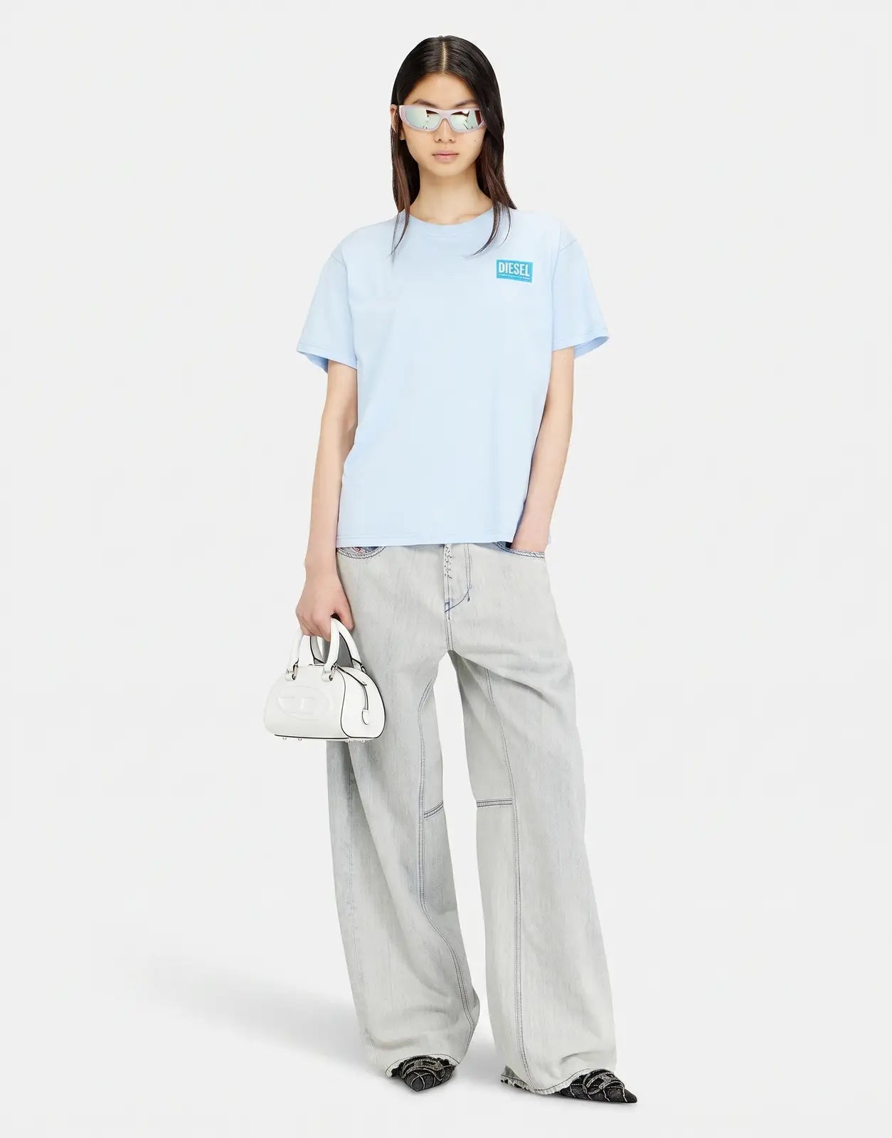Female model wears a light blue Diesel T-Normy S6 Regular T-shirt, light wash wide-leg jeans, and holds a white handbag.