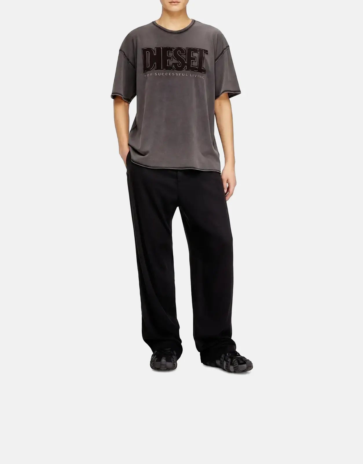 Male model wearing a dark grey Diesel T-Rawboxt relaxed t-shirt, black pants, and a distressed cap, standing full body.