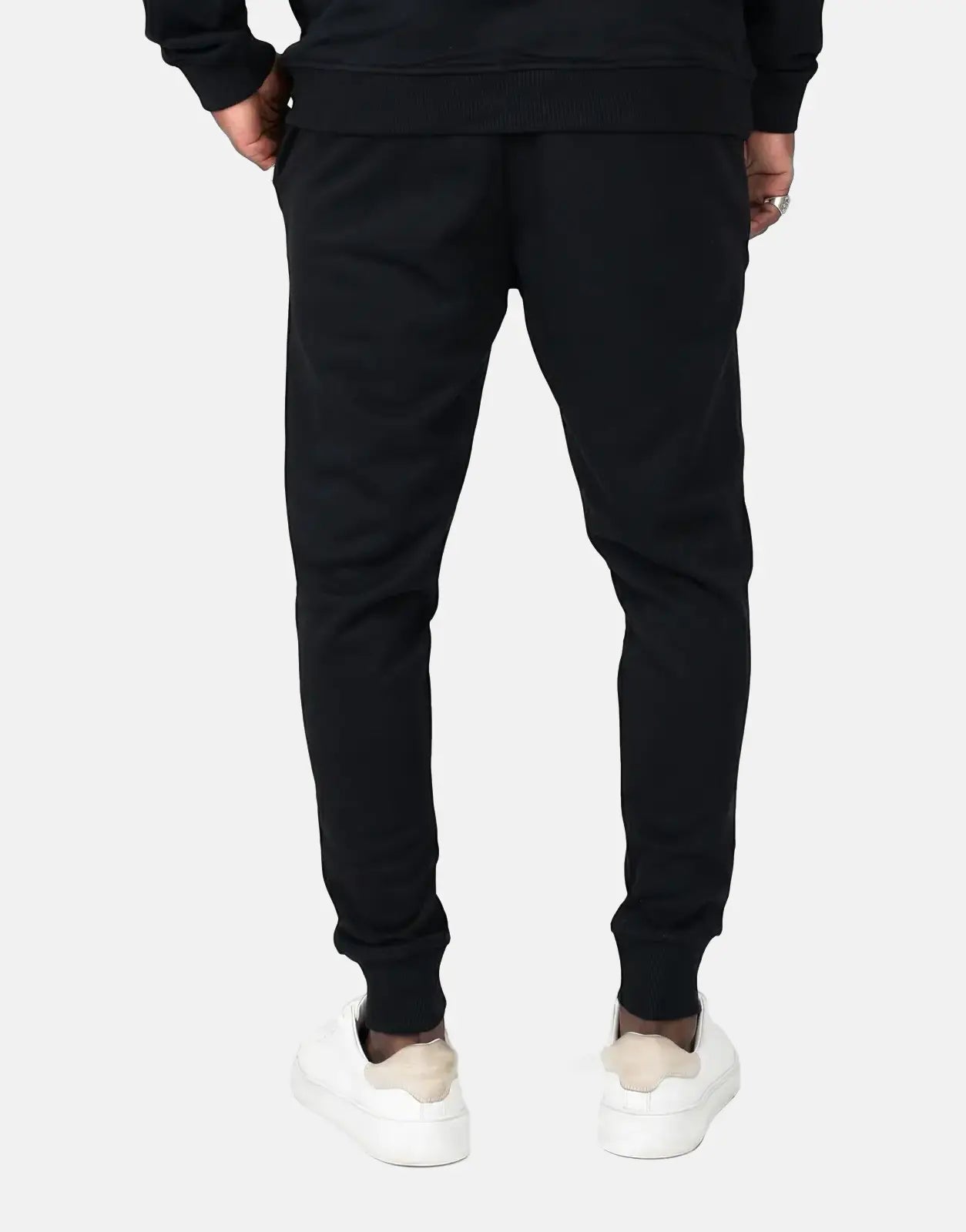 A person from the back wearing black Fade Signature sweatpants with ribbed cuffs and white sneakers on a simple background.