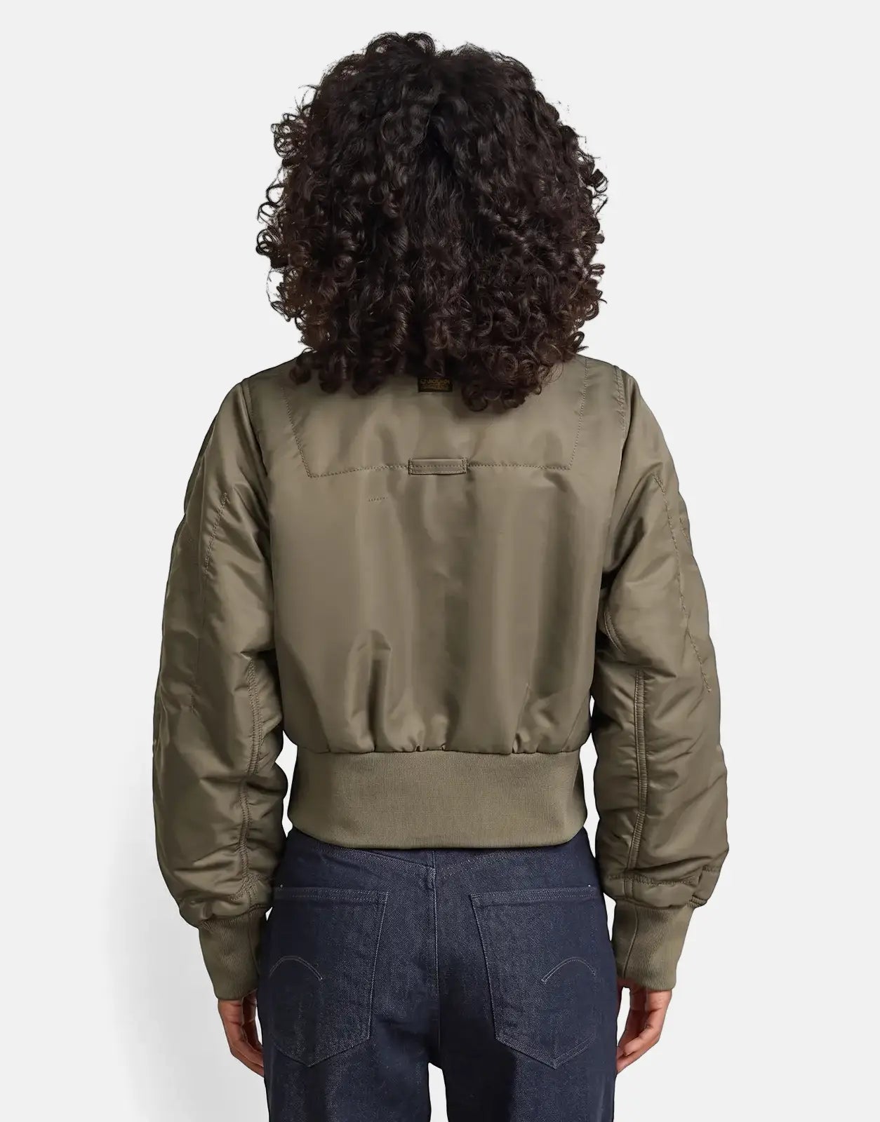 A woman from behind models an olive green G-Star RAW Bomber Turf jacket and dark jeans, showcasing the jacket's ribbed
