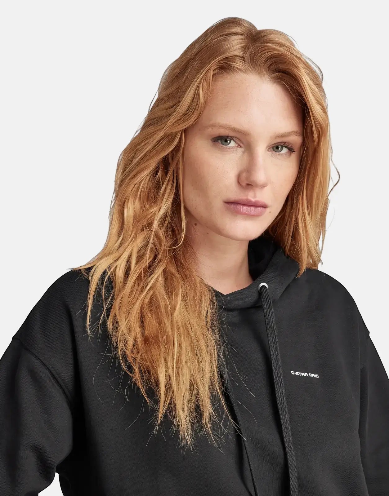 Woman with reddish-blonde hair wearing a black G-Star RAW Graphic Core Hood. Studio close-up of the pullover hoodie.