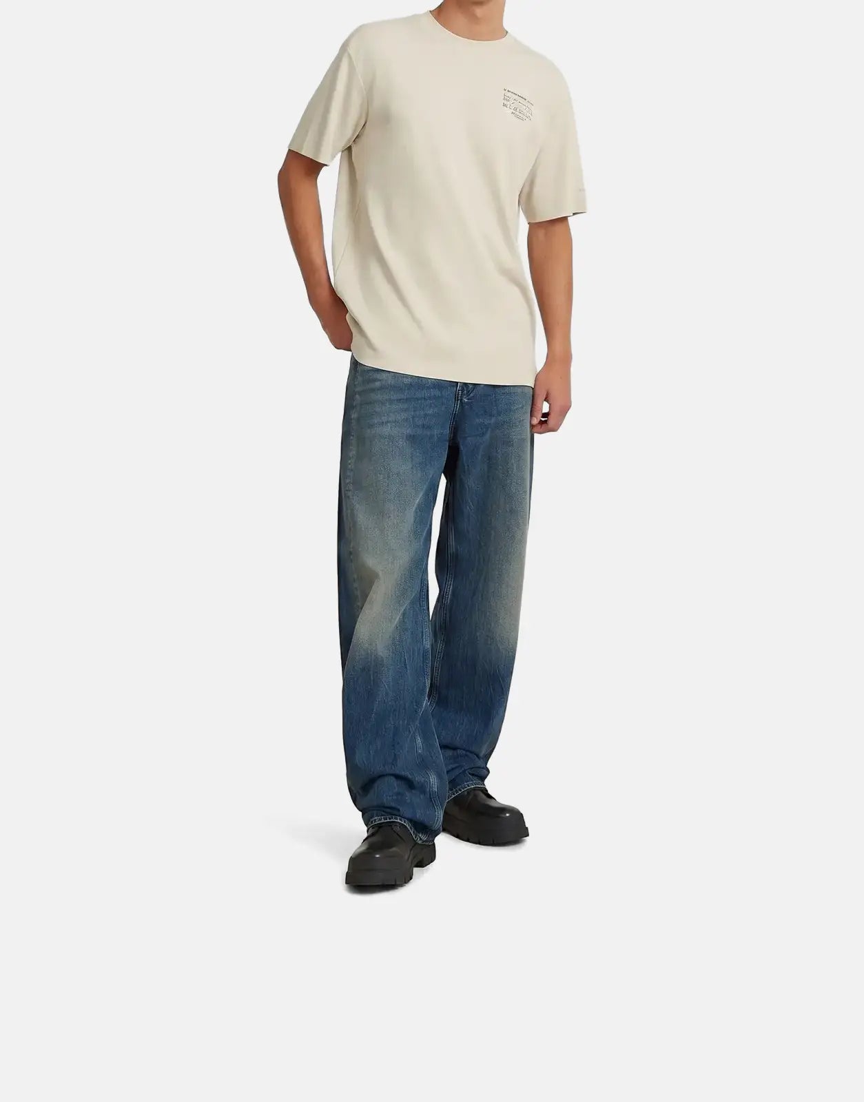 A male model wears a G-Star RAW Script Whitebait T-Shirt in cream with blue baggy jeans and black chunky shoes.