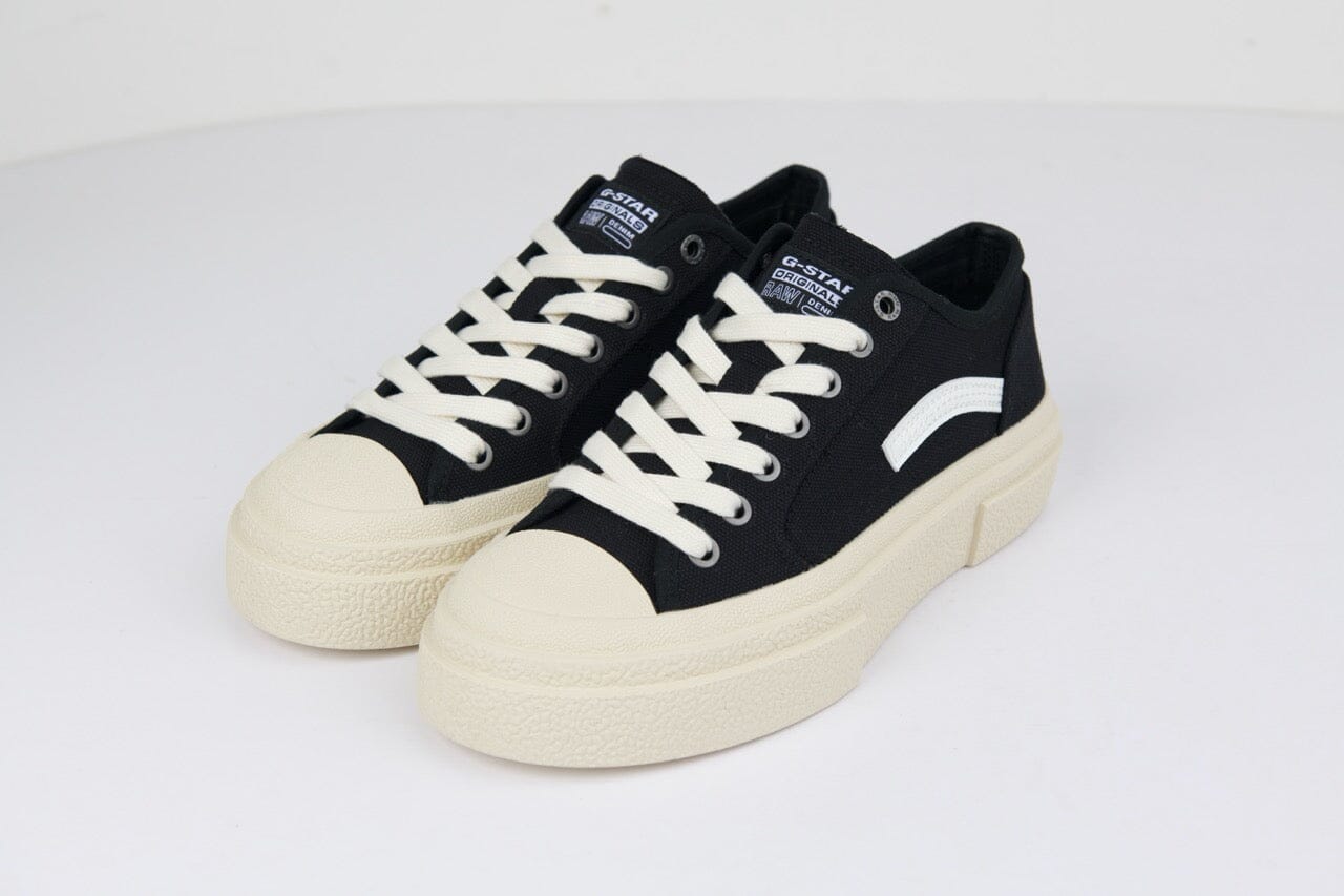 Black canvas sneakers with a cream rubber toe cap, beige laces, and subtle white branding on the side. Versatile casual footwear.