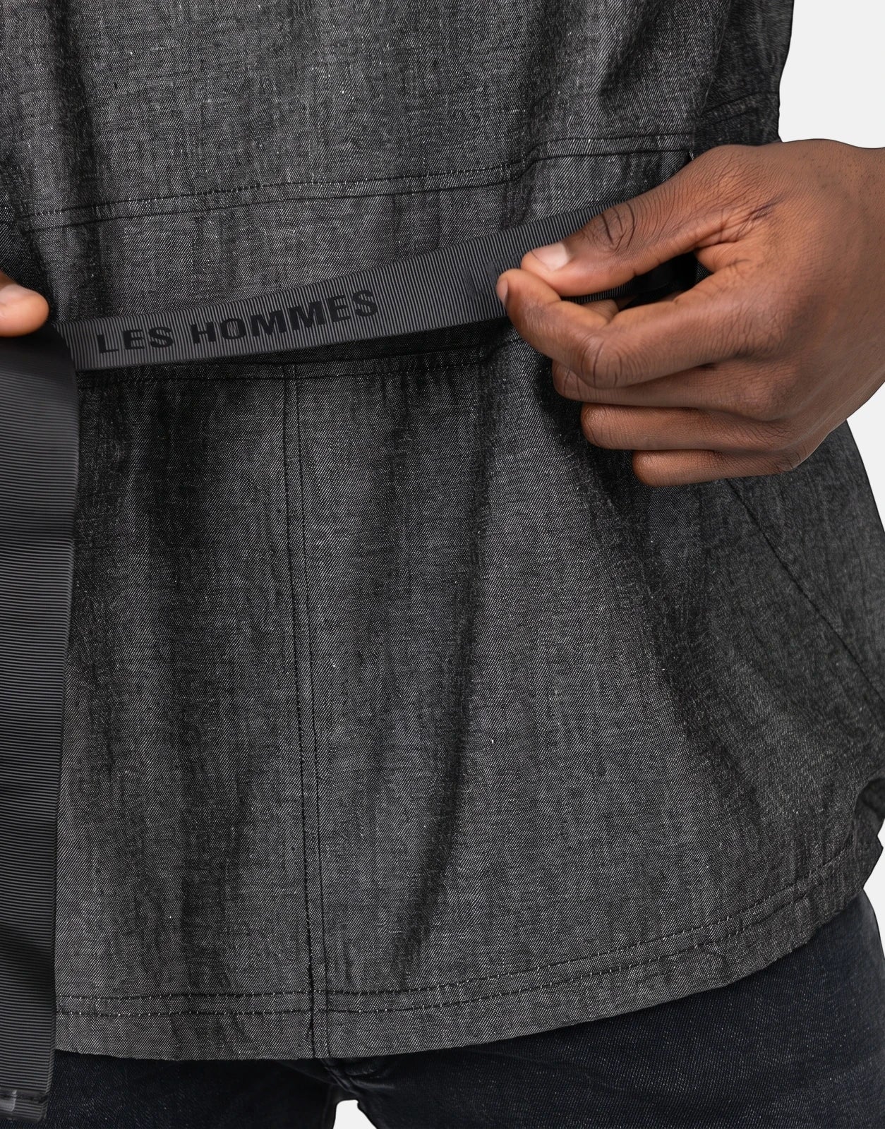Close-up of a person wearing a dark grey Les Hommes t-shirt, with hands adjusting the black ribbed belt.