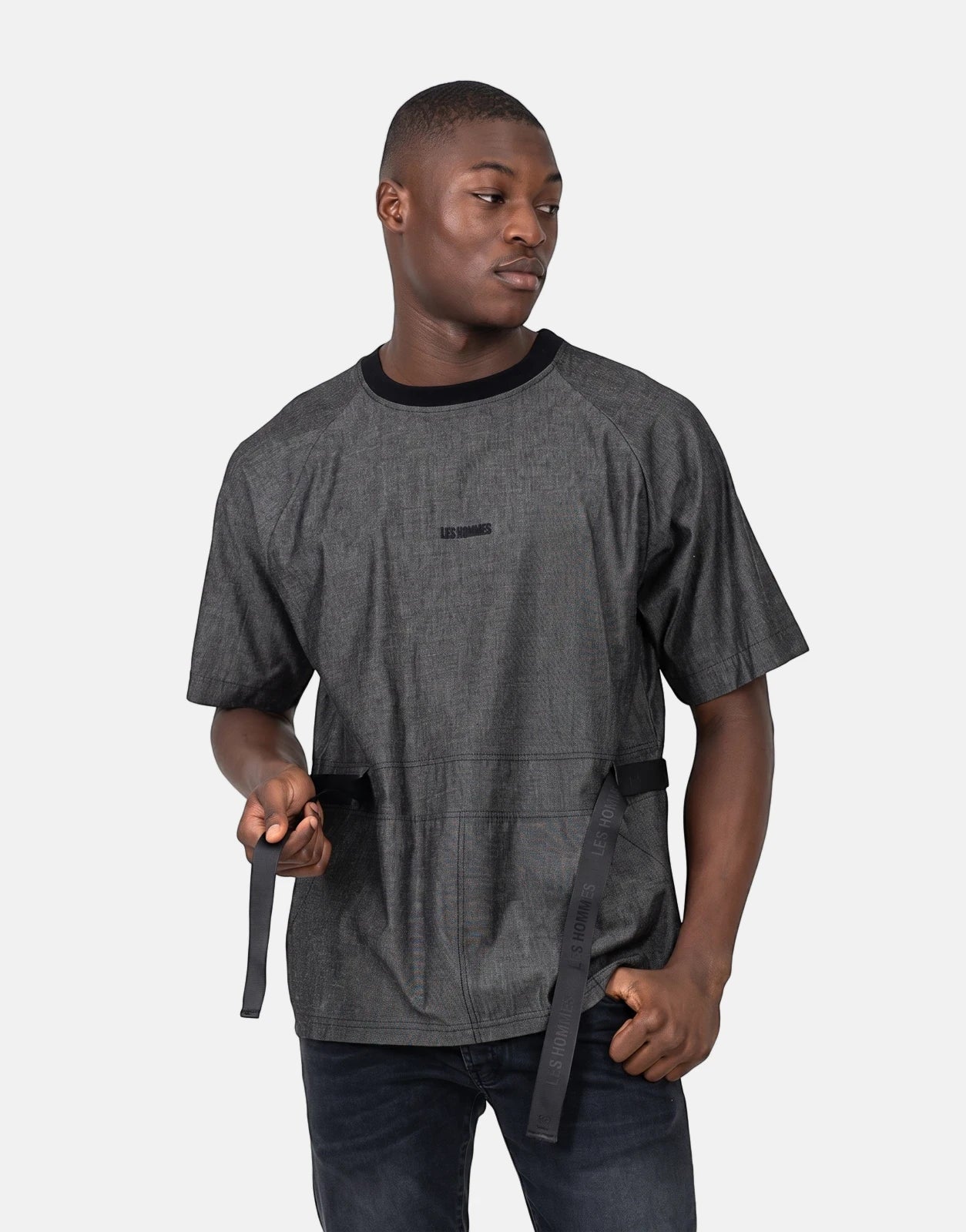 A male model wears a Les Hommes grey t-shirt with distinctive side strap detail and dark jeans, shown in a front studio view.