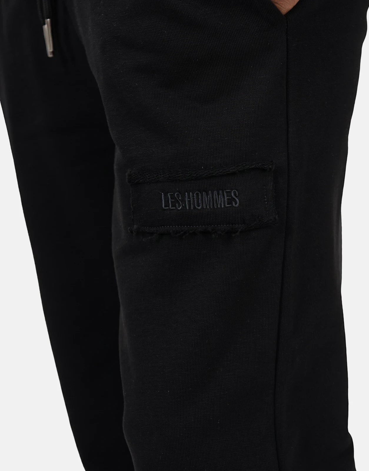 Close-up of Les Hommes black jogger pants featuring a stitched logo label and a silver drawstring tip, side view.