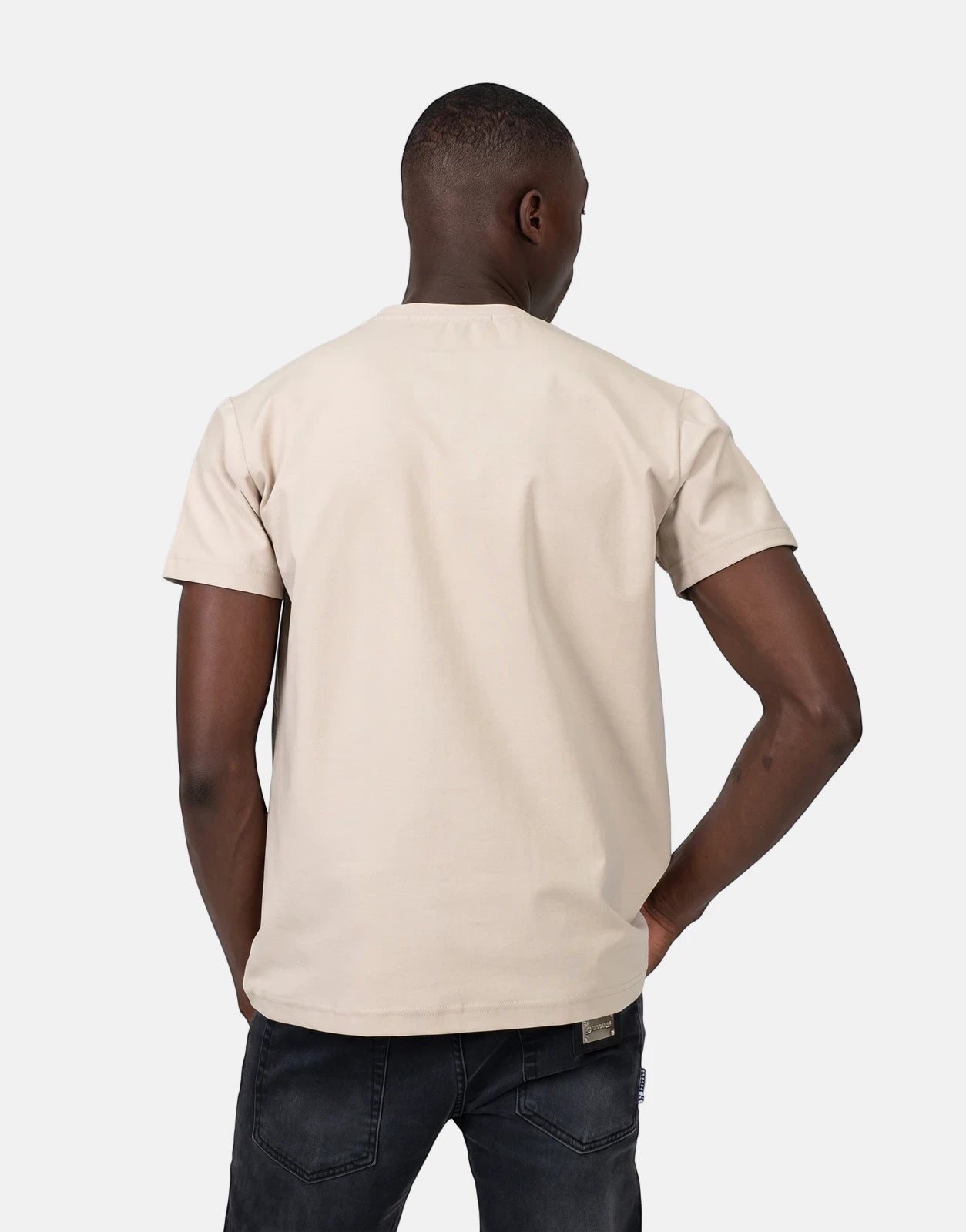 Back view of a male model wearing a brown Les Hommes t-shirt with short sleeves and black jeans, standing against a simple