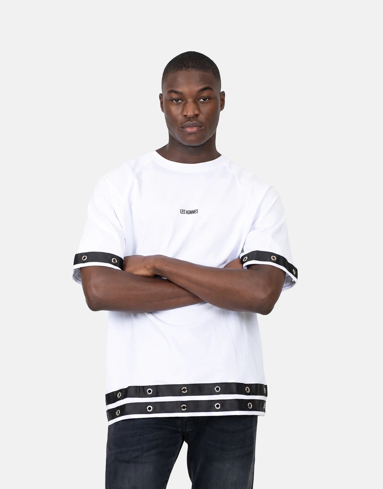 A male model poses in a Les Hommes Denim Insert White T-Shirt featuring black eyelet details on the sleeves and hem.