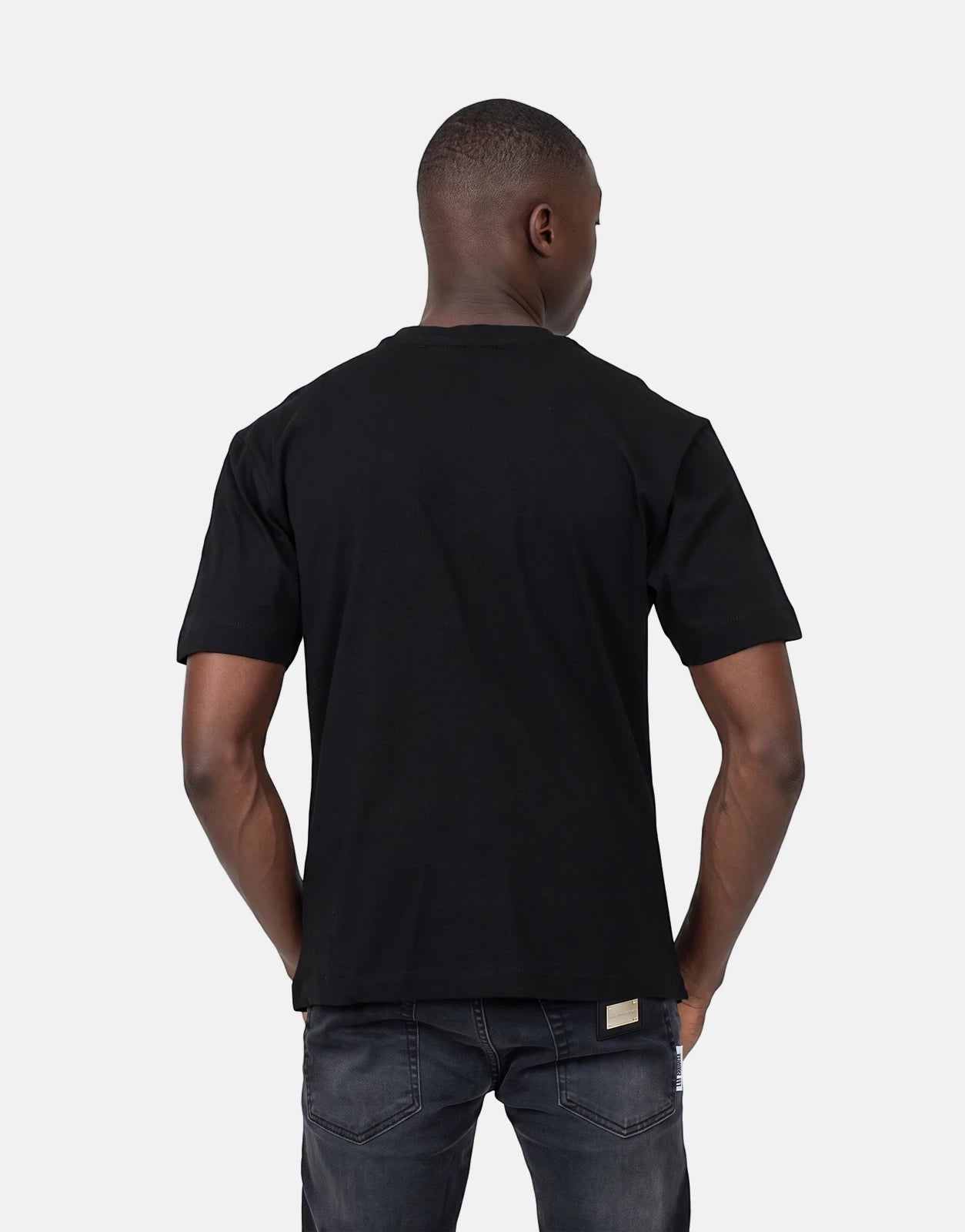 Back view of a man wearing a black Les Hommes Dragonfly T-Shirt and dark wash jeans, standing with hands in pockets.