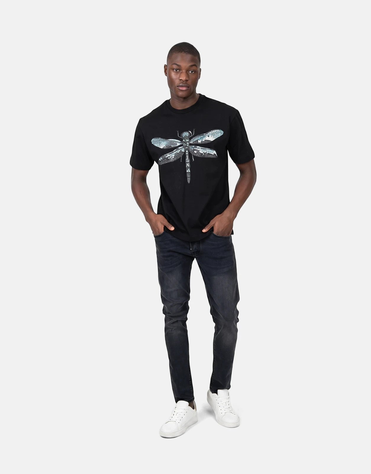 A male model wears a black Les Hommes Dragonfly T-shirt, dark wash jeans, and white sneakers, standing full-body.