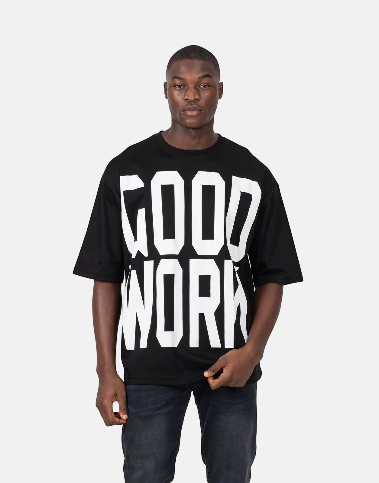 A man models a black Les Hommes Good Work T-shirt, featuring bold white text on the front, paired with dark jeans.