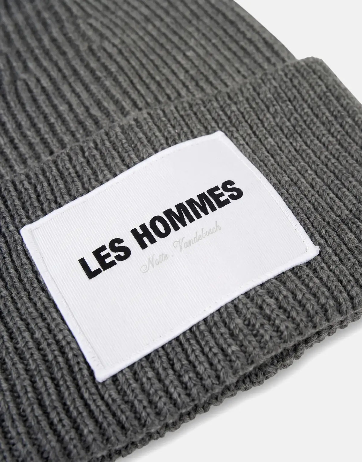 Close-up of a grey Les Hommes ribbed beanie featuring a white logo label with black text, positioned on a white surface.