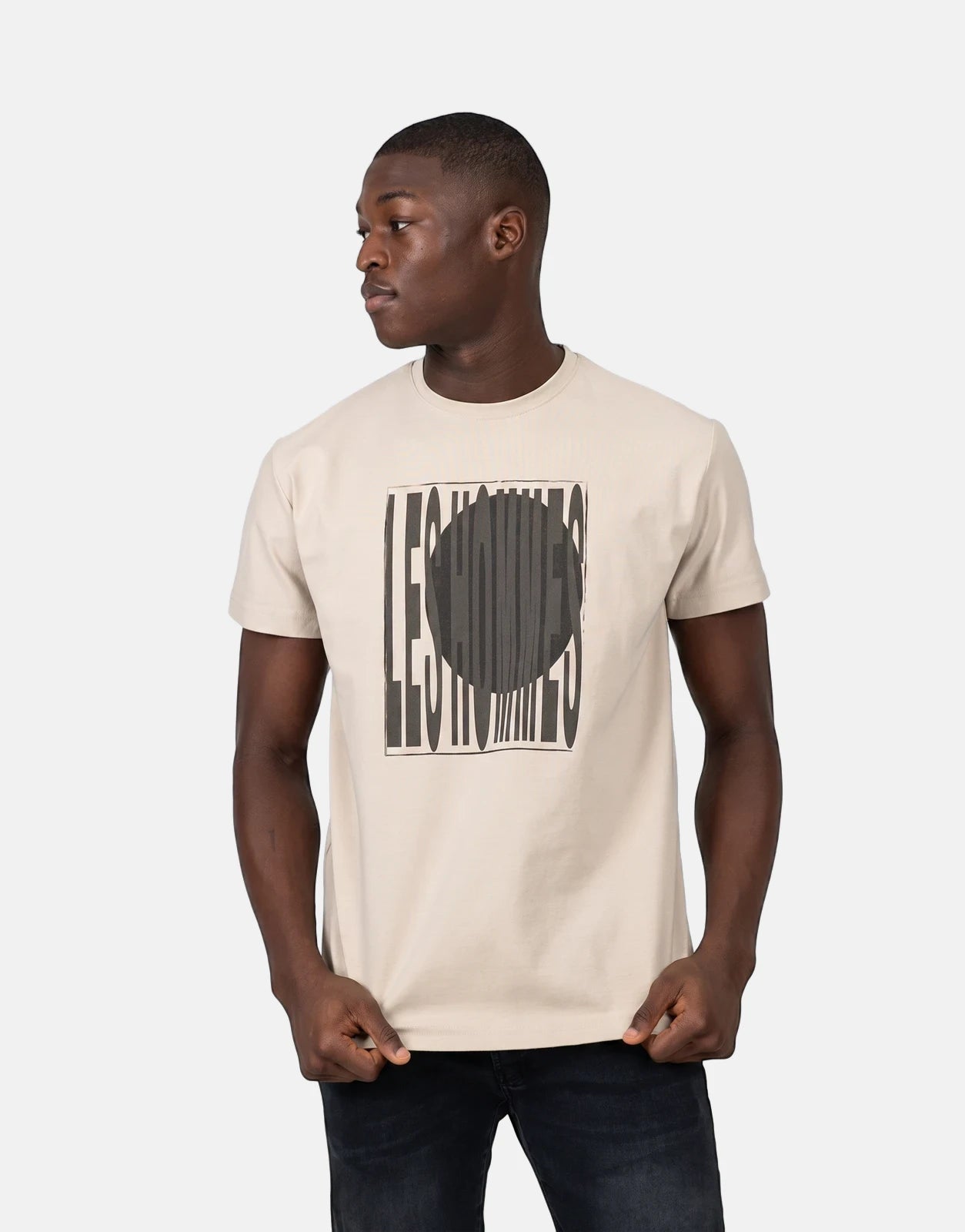 Male model wearing a beige Les Hommes Logo Print T-Shirt with a black graphic, paired with dark jeans.