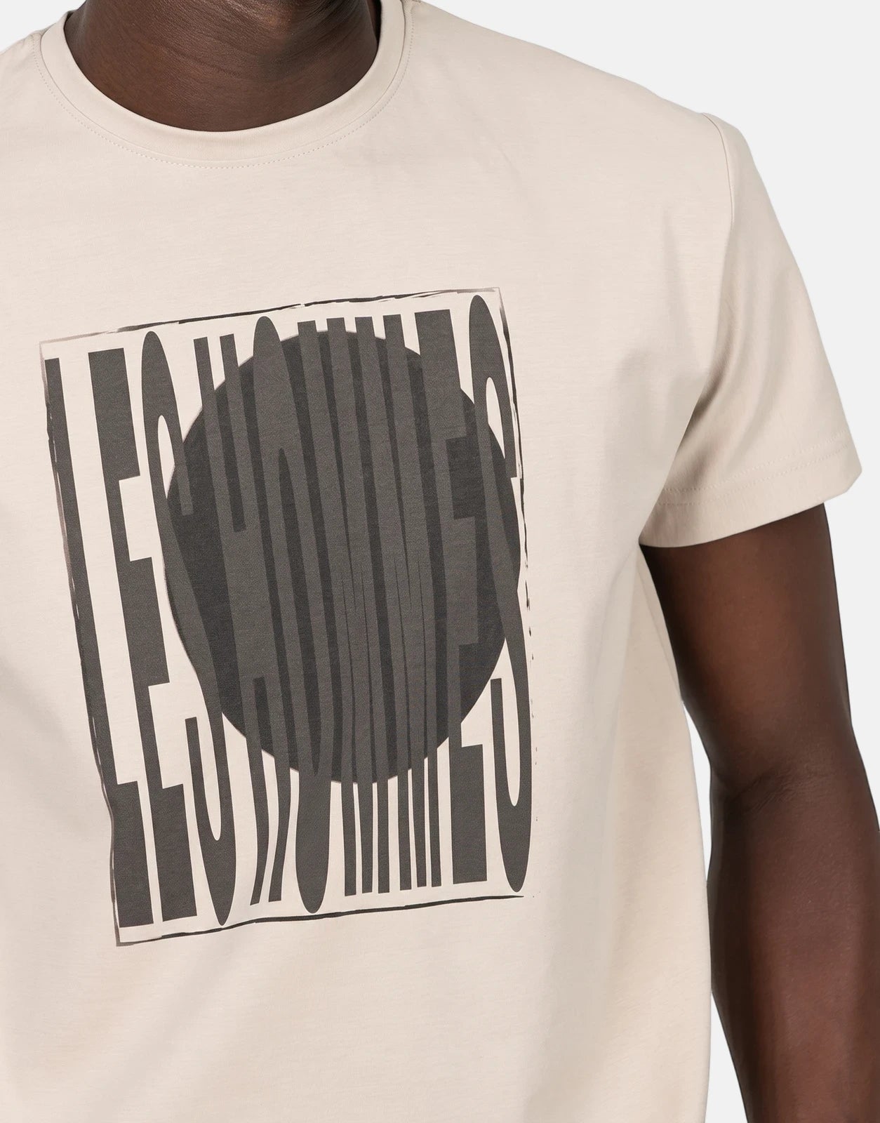Close-up of a person wearing a Les Hommes Logo Print Brown T-Shirt, featuring a distinct black and white graphic design.