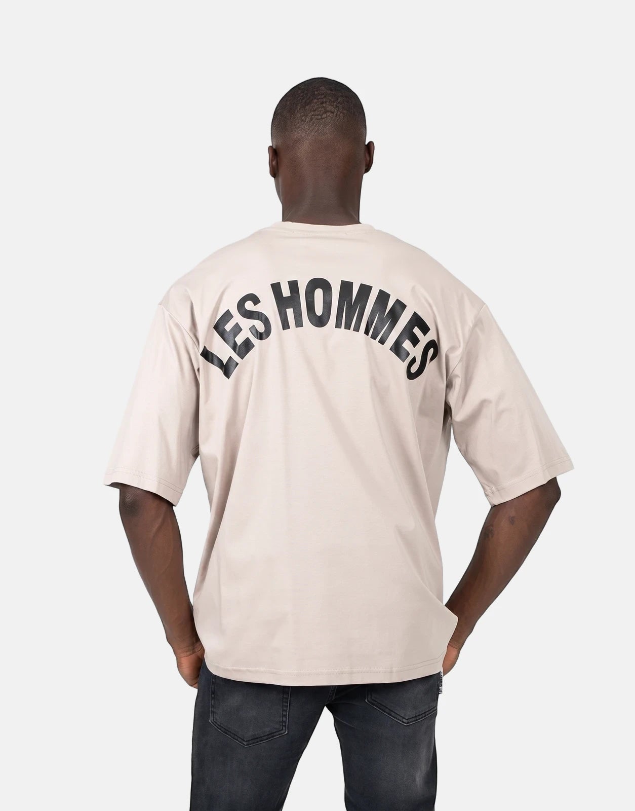 Male model from back wearing a Les Hommes Over Logo Back brown t-shirt with large black lettering on a plain background.