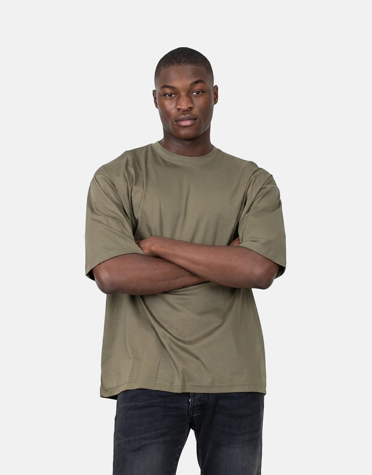 Young man wearing an olive green Les Hommes Over Logo Back T-Shirt and black jeans with arms crossed, photographed from the