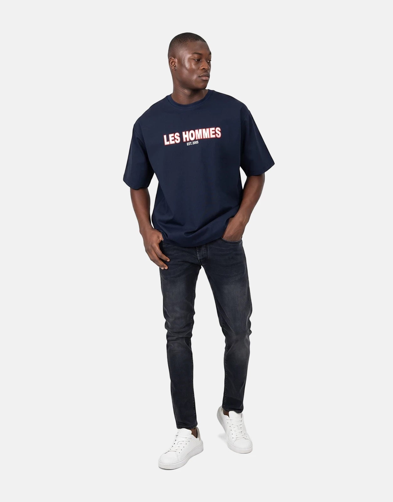 Male model wearing a Les Hommes Over Rubber Print Blue T-Shirt with black jeans and white sneakers, full body shot.