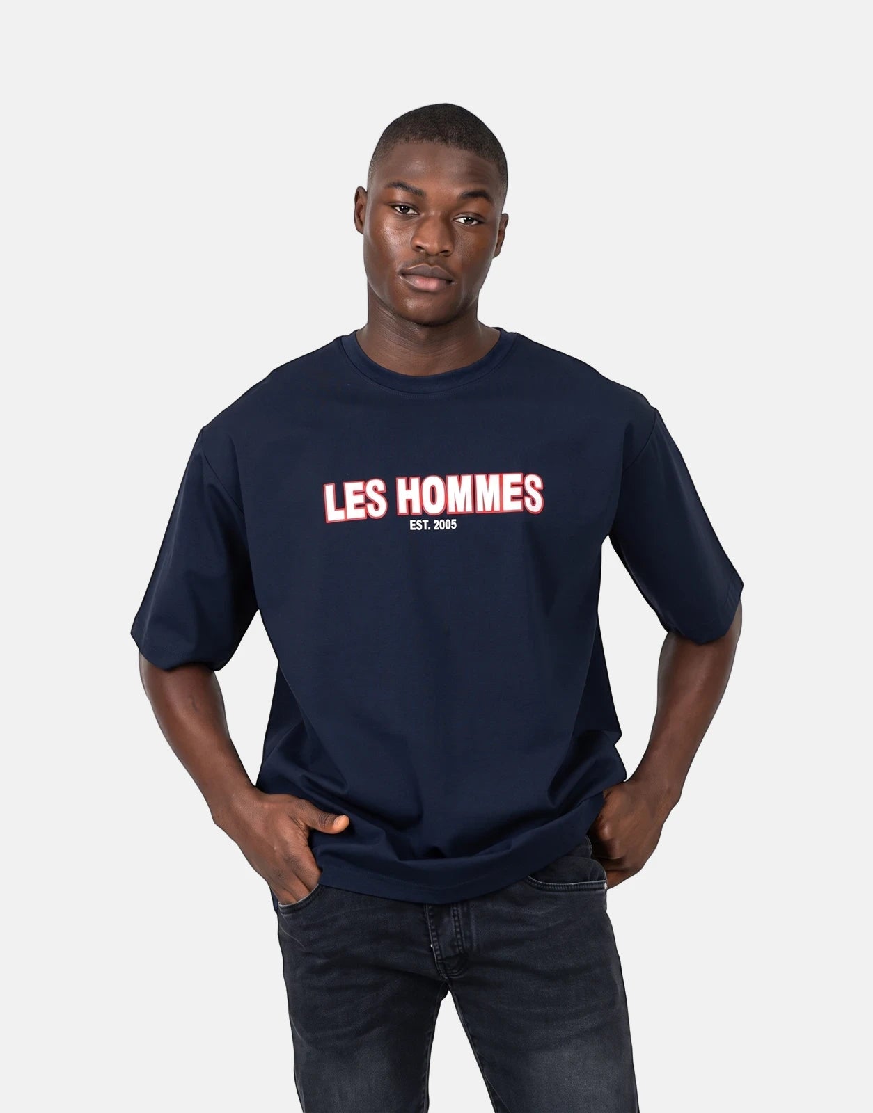 Male model wearing a navy blue Les Hommes Over Rubber Print T-Shirt with a white and red logo, paired with dark jeans.