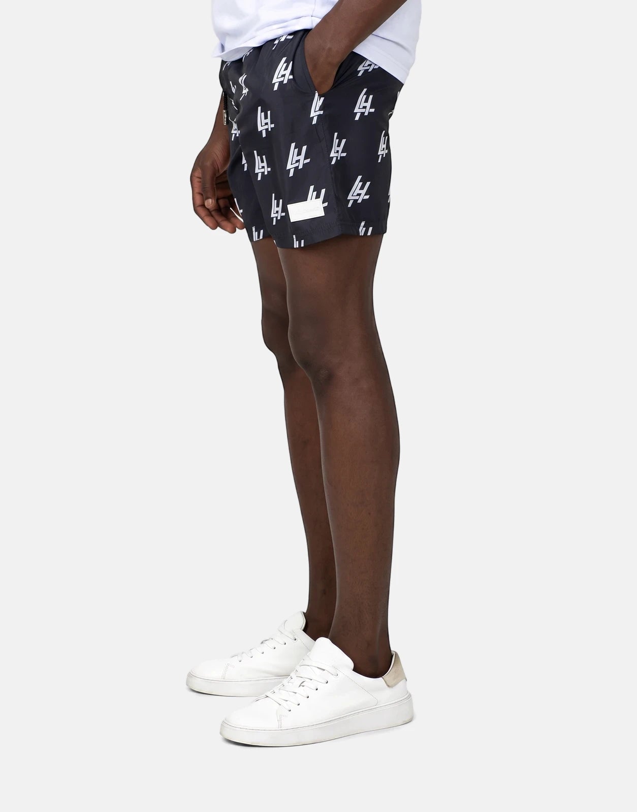 Les Hommes Print Black Swim Shorts with white LH monogram pattern worn by a model, captured in a side profile view.