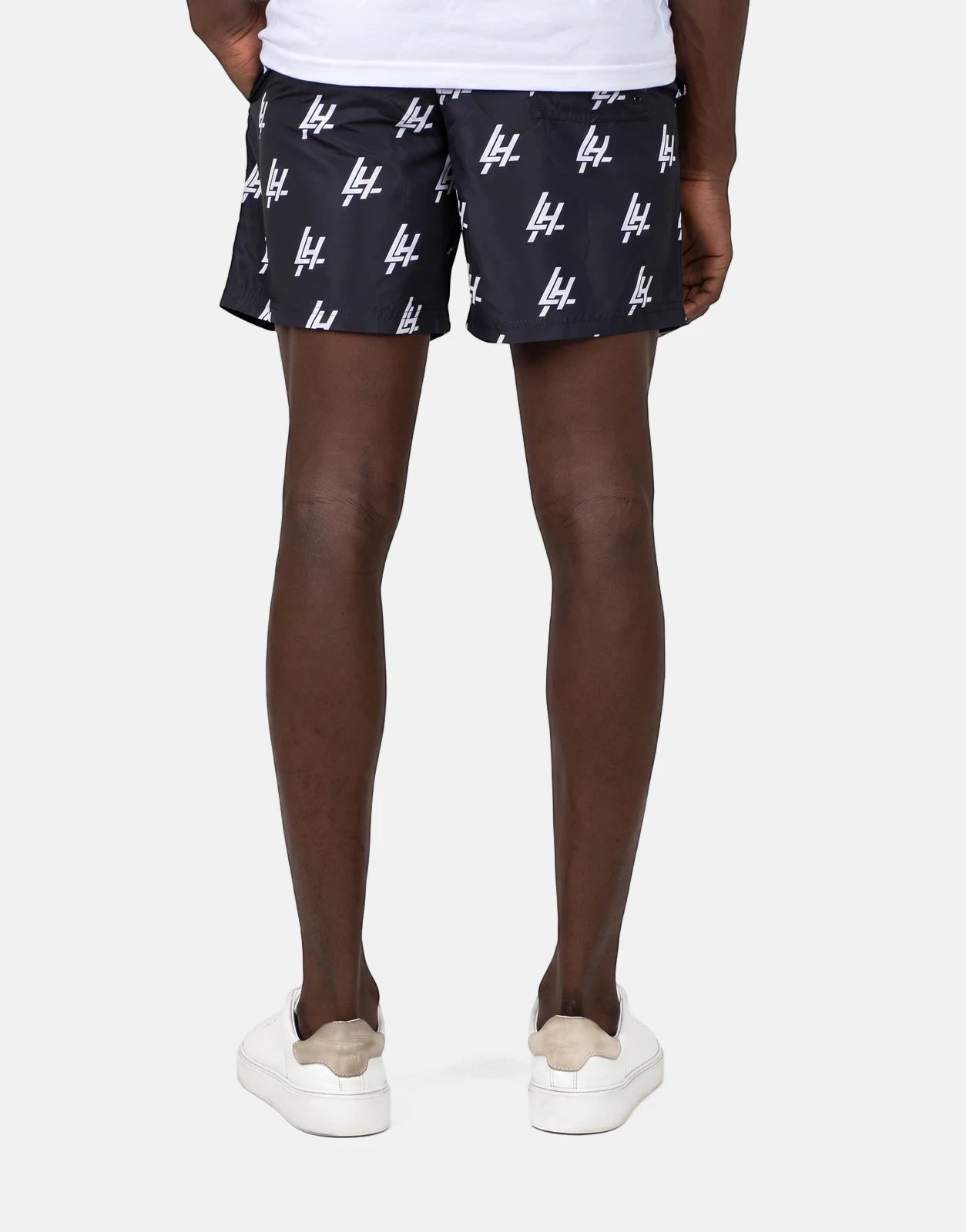 A person's lower body from behind, wearing black Les Hommes swim shorts with white logo print and white sneakers.