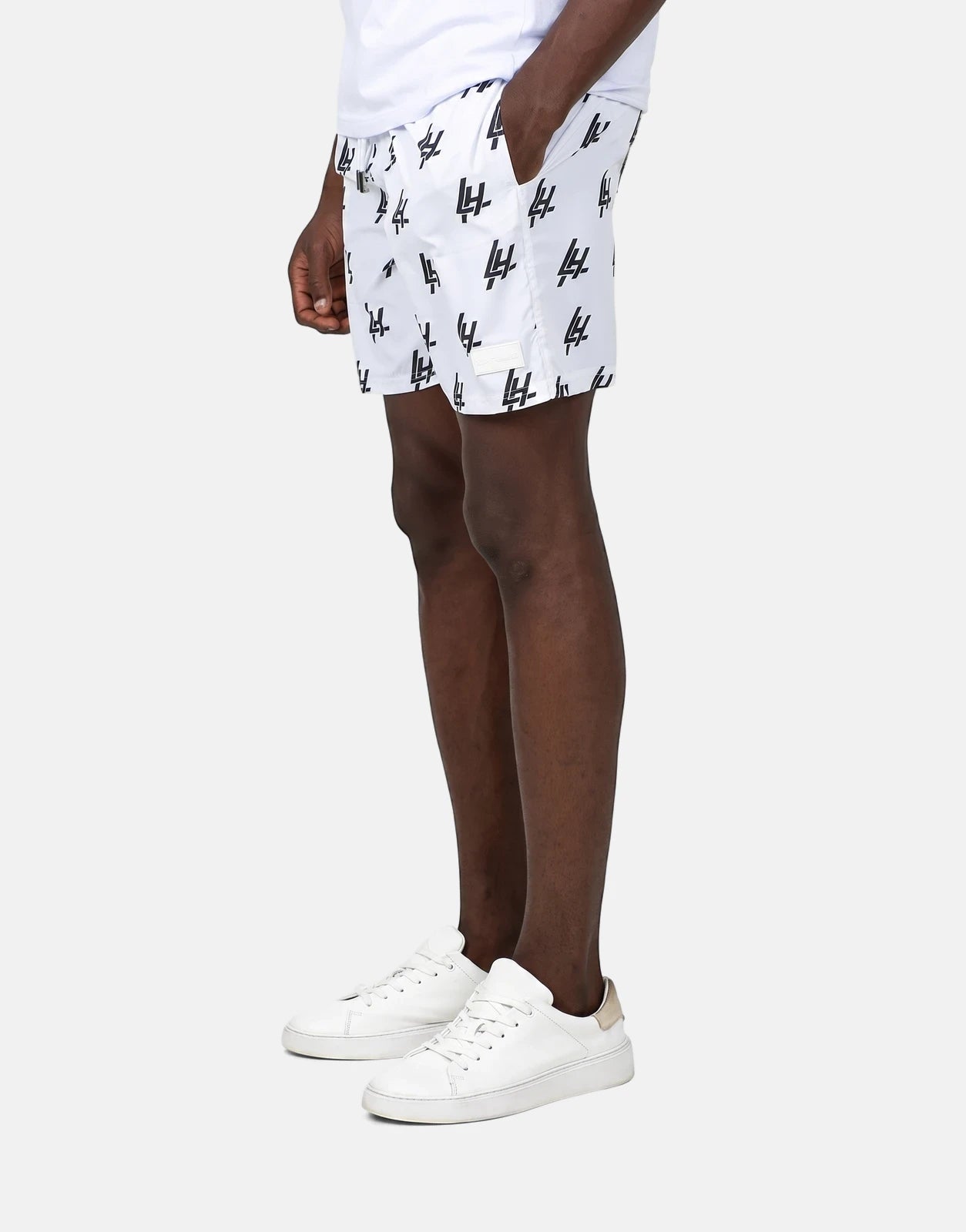 Les Hommes white swim shorts with a black 'LH' print, worn by a male model in a side view, paired with white sneakers.