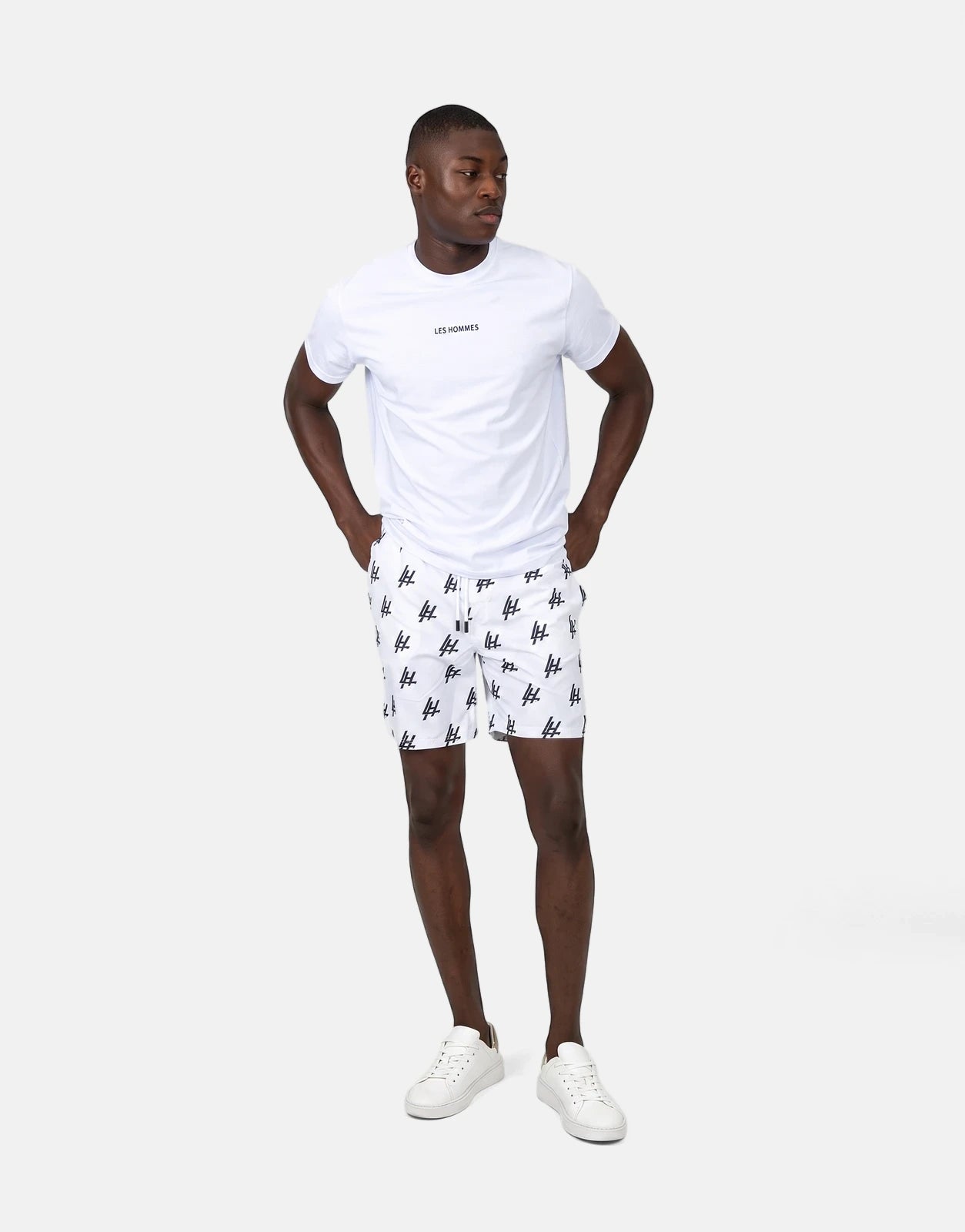 Male model in Les Hommes white swim shorts with black monogram print and a white t-shirt, full body view.