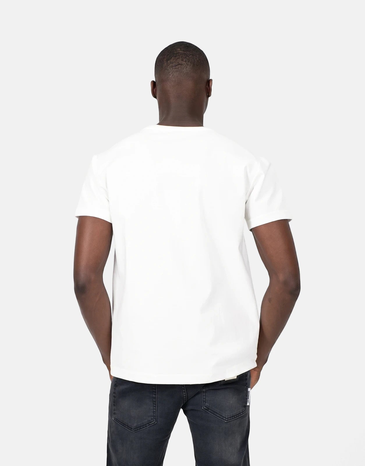 Back view of a man wearing a plain white short-sleeve crew neck t-shirt and dark wash jeans, standing casually.