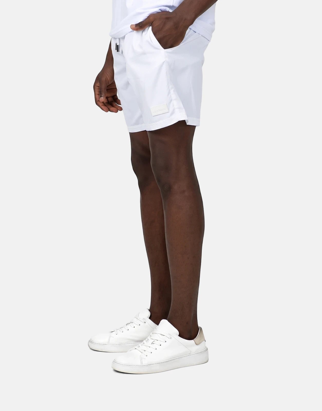 Male model showcasing Les Hommes white swim shorts, paired with white sneakers, in a side profile studio shot.