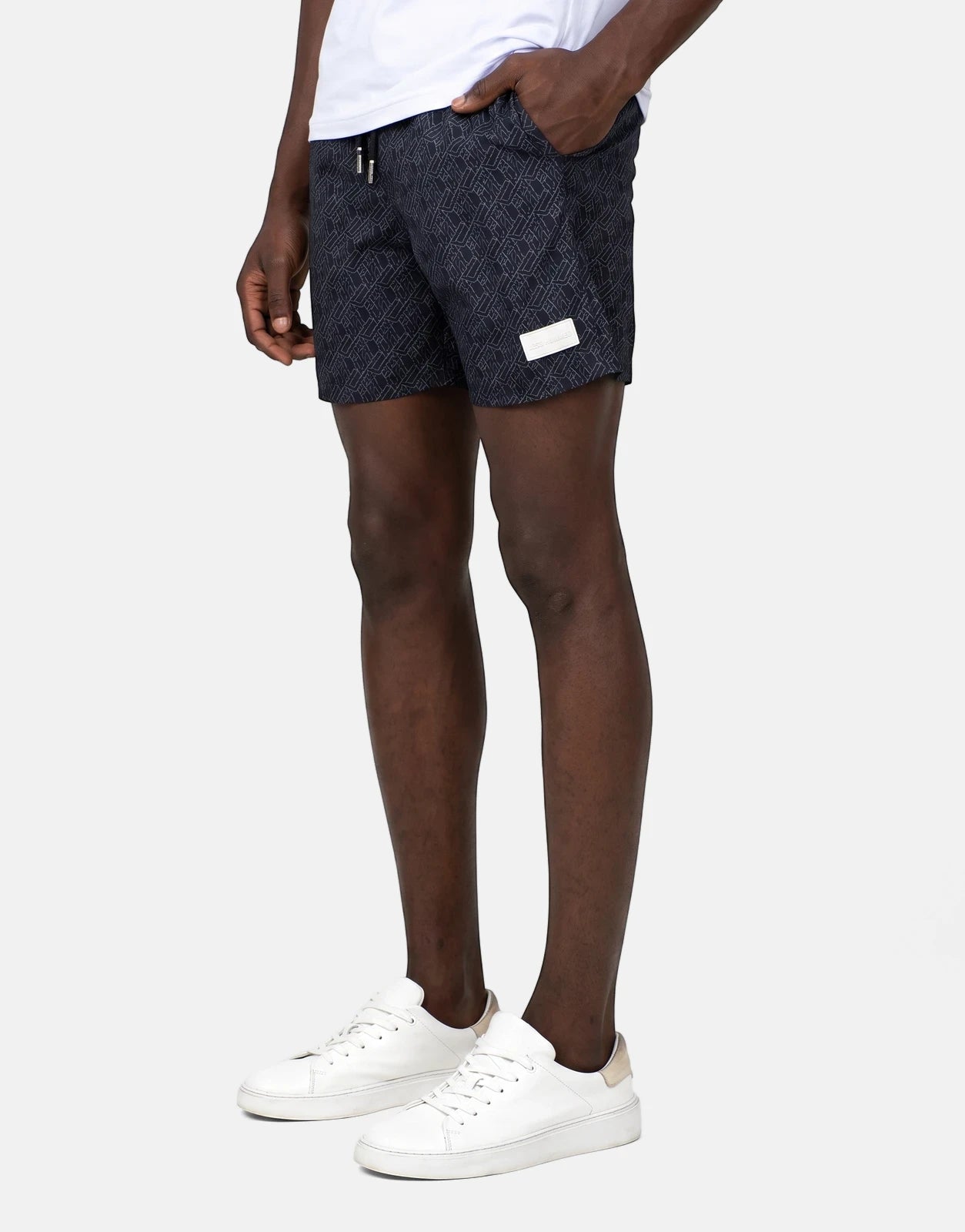 Men's Less Hommes AOP black swim shorts with a geometric pattern, worn by a model with white sneakers.