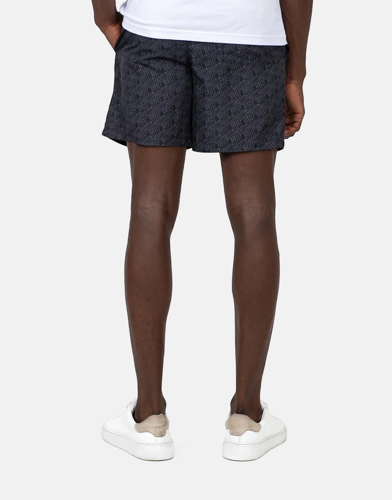 Back view of a person wearing Less Hommes AOP black patterned swim shorts with white sneakers on a simple background.