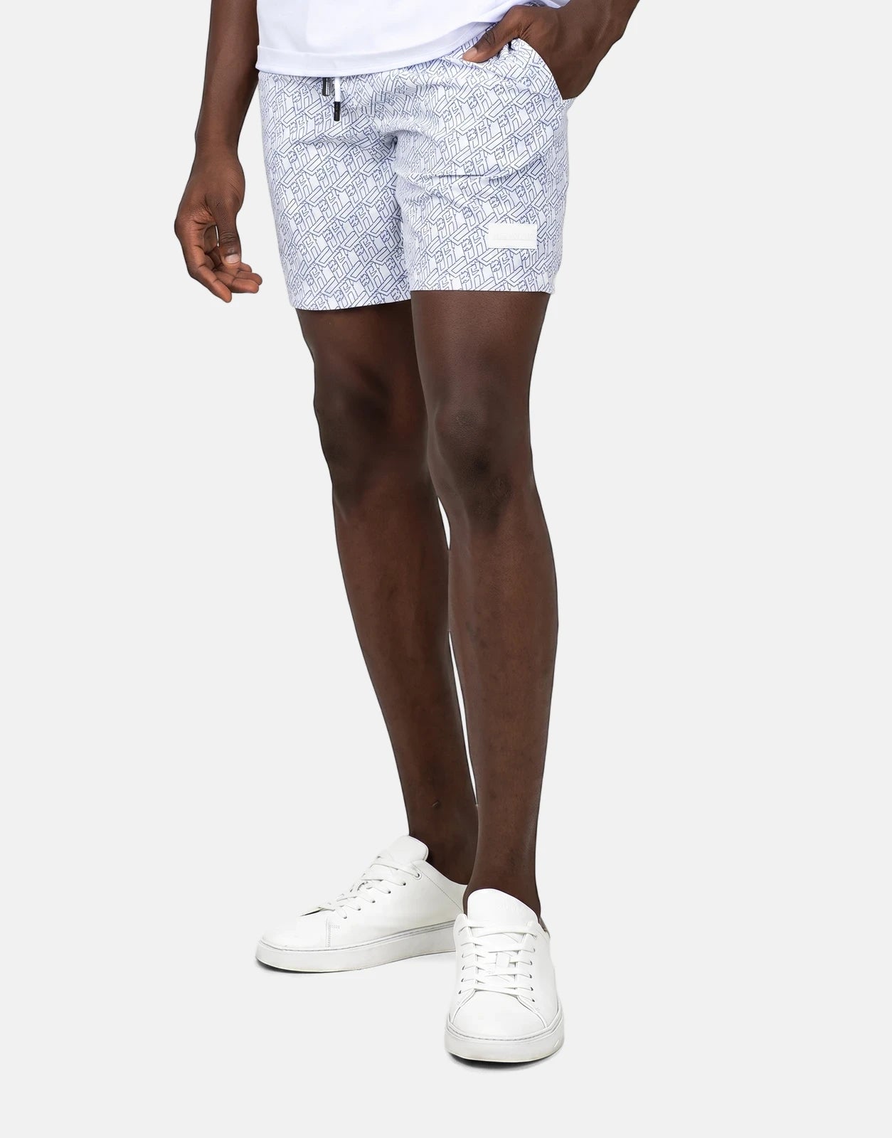 Men's Less Hommes AOP white swim shorts with a distinctive monogram pattern, worn by a model standing.