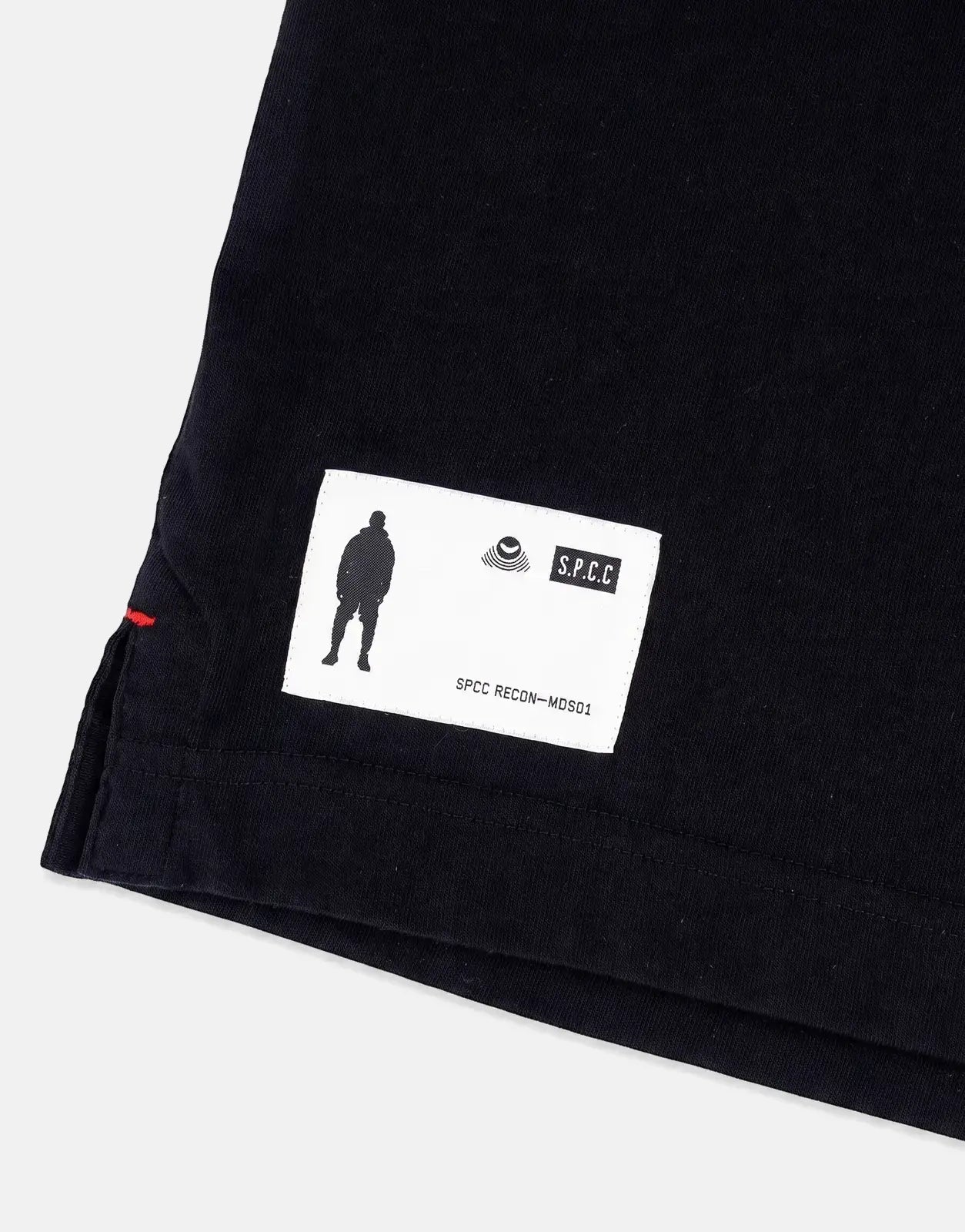 Close-up of the bottom corner of an SPCC Recon Overwatch black t-shirt, showing the white brand label and red side stitching.