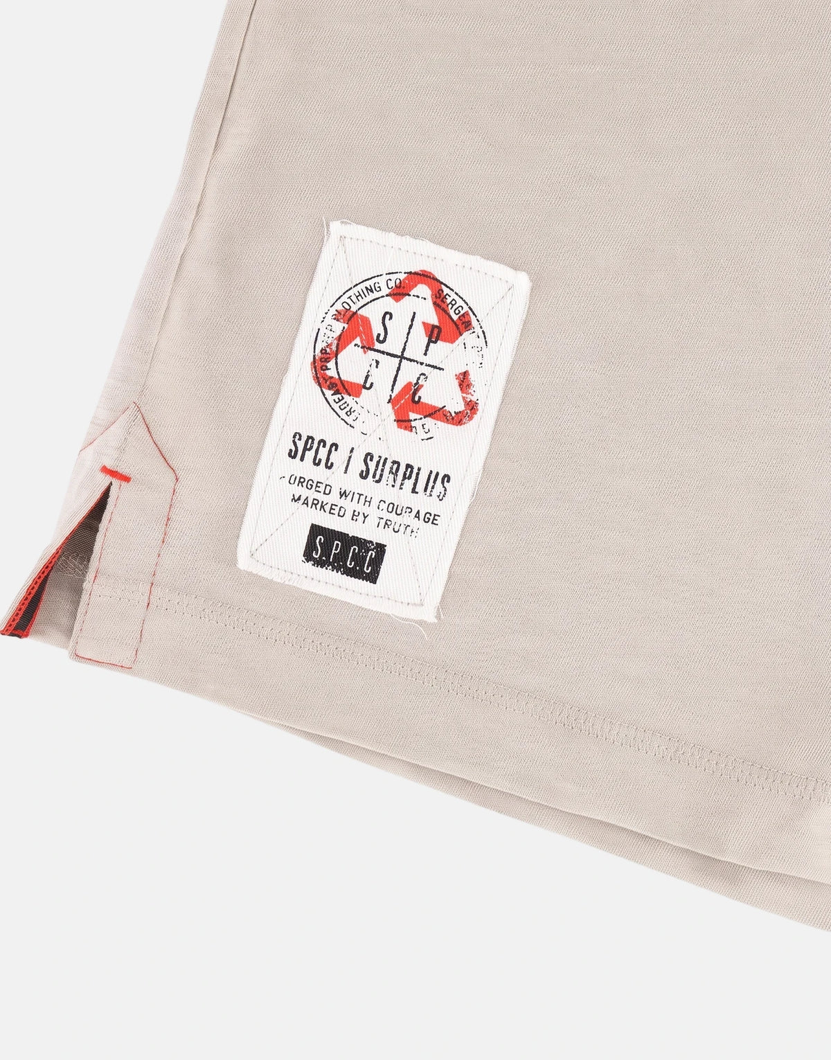 A close-up of a beige SPCC Troy tee featuring a distressed fabric label with red and black graphics and red stitched detail