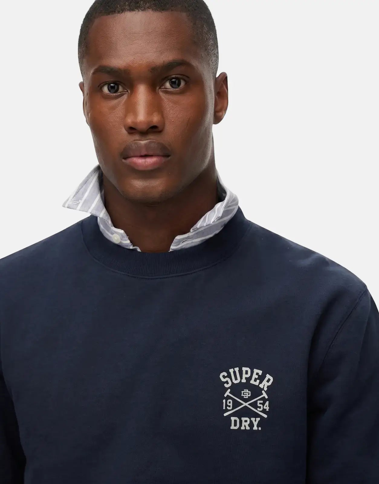 Close-up of a man wearing a navy blue Superdry Athletic Club sweatshirt with a white logo, layered over a striped collared