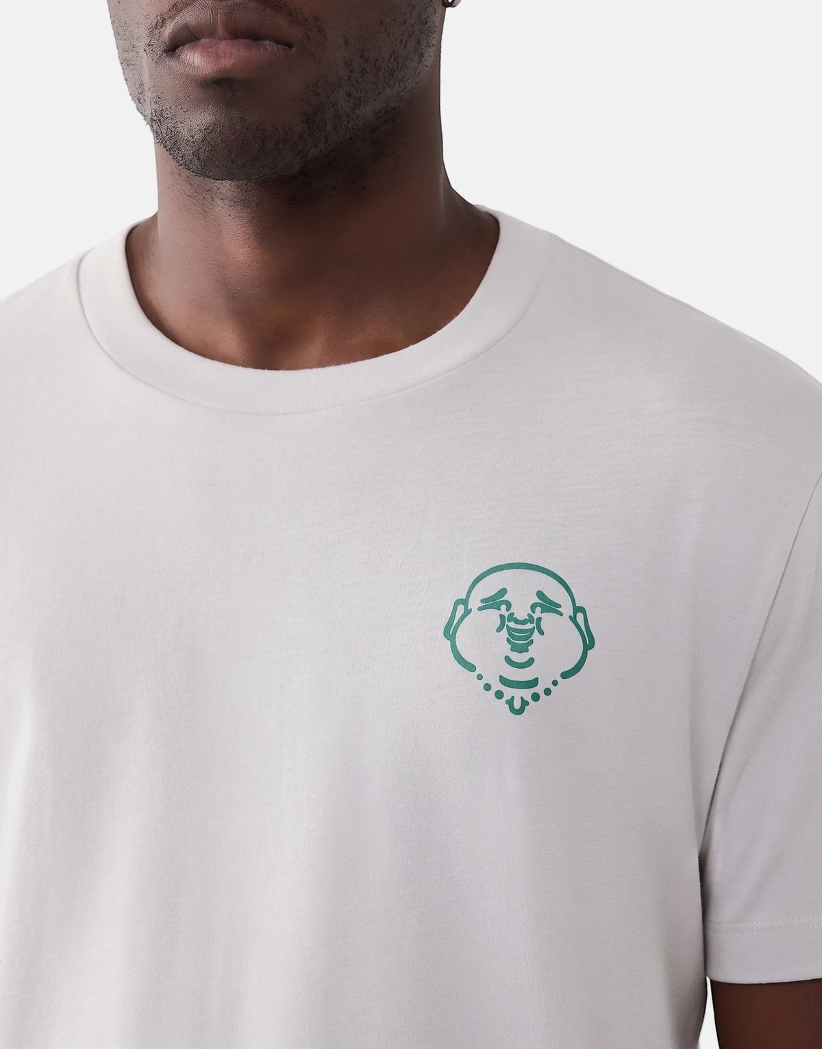 Close-up of a man wearing a light grey True Religion Abstract Denim T-Shirt with a green Buddha logo on the chest.