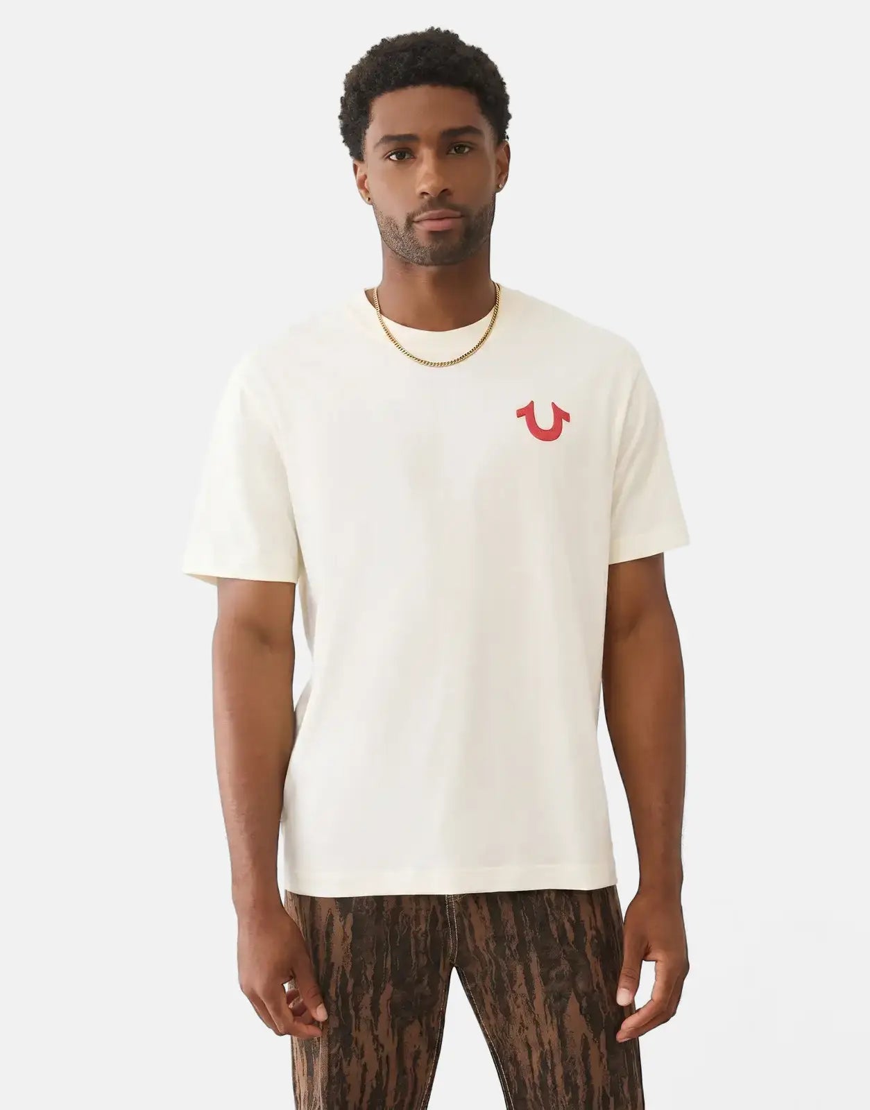Male model wearing a cream True Religion Number One SRS T-shirt with a red horseshoe logo and textured brown pants.
