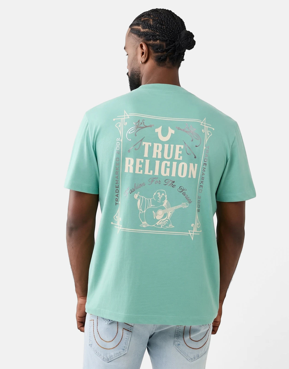 Male model from behind wearing a True Religion Rect Beryl Green T-shirt with a graphic back print and light wash denim jeans.